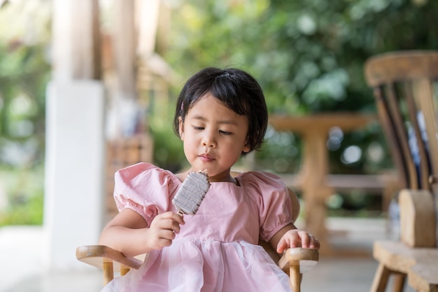 Foco raso de uma jovem garota do Sudeste Asiático comendo um picolé enquanto está sentado em uma cadeira de madeira