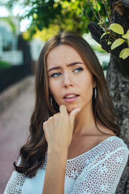 Foco superficial de uma mulher morena vestindo uma camisa de renda branca e fazendo uma careta pensativa em um parque