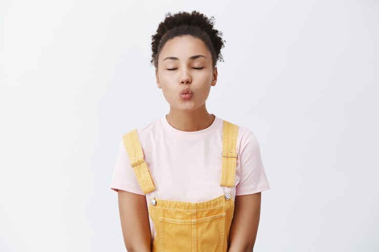 Foto grátis fofa mulher afro-americana em macacão amarelo, dobrando os lábios para fazer mwah, fechando os olhos e parada apaixonadamente sobre uma parede cinza
