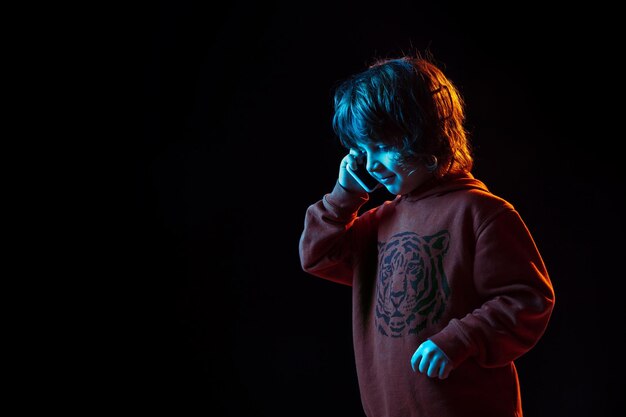 Fofo falando no telefone. Retrato do menino caucasiano em fundo escuro do estúdio em luz de néon. Lindo modelo cacheado.