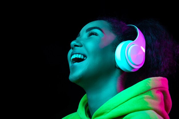 Fones de ouvido. Retrato de uma mulher afro-americana isolado no fundo escuro do estúdio em luz de néon multicolorida. Lindo modelo feminino. Conceito de emoções humanas, expressão facial, vendas, anúncio, moda.