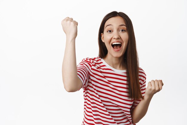 Força equipa. Menina morena animada torcendo, levantando o punho cerrado, ativista torcendo e cantando, sorrindo esperançoso, comemorando a vitória, de pé contra um fundo branco