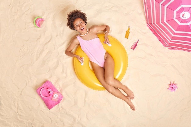 Foto aérea de mulher de cabelo encaracolado feliz em poses de roupa de banho na natação inflada amarela passa o tempo livre na praia encontra-se no sol cercado por brinquedos de areia garrafa de bebidas energéticas fones de ouvido na toalha