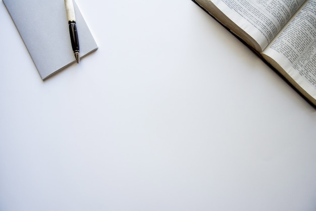 Foto aérea de uma Bíblia aberta e um bloco de notas com uma caneta em uma superfície branca