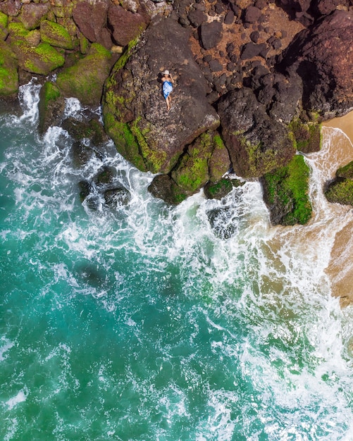 Foto aérea vertical de uma mulher com um vestido azul deitada na praia rochosa