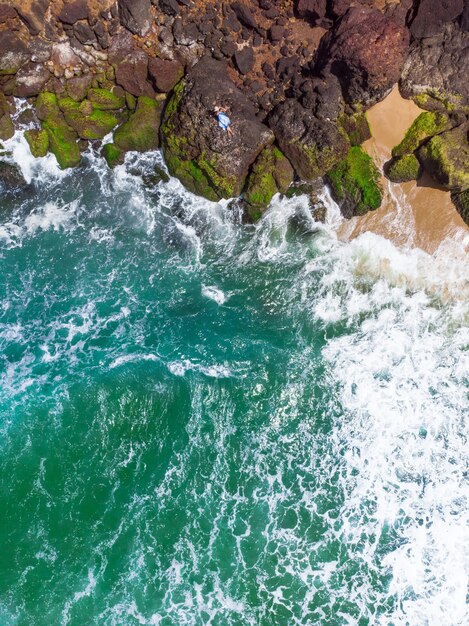 Foto aérea vertical de uma mulher com um vestido azul deitada na praia rochosa