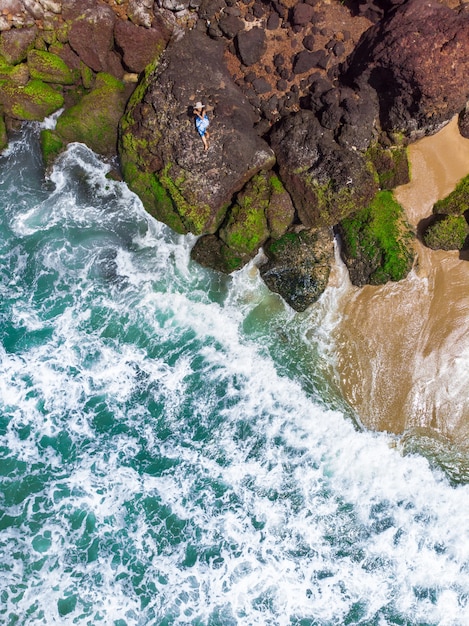 Foto aérea vertical de uma mulher com vestido azul deitada na praia rochosa