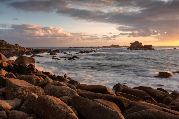 Foto ampla do oceano com formações rochosas na costa durante o pôr do sol com céu nublado
