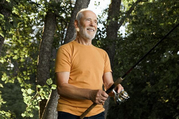 Foto ao ar livre de atraente homem caucasiano sênior com barba por fazer segurando uma vara de fiar nas águas do rio, sorrindo com antecipação, esperando o peixe ser fisgado, o sol brilhar e as árvores verdes em