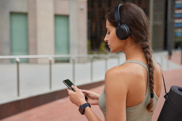 Foto ao ar livre de mulher esportiva com rabo de cavalo usa notificação de verificação de telefone celular ouve faixa de áudio via fones de ouvido sem fio carrega suportes de karemat enrolados contra fundo desfocado estilo de vida ativo