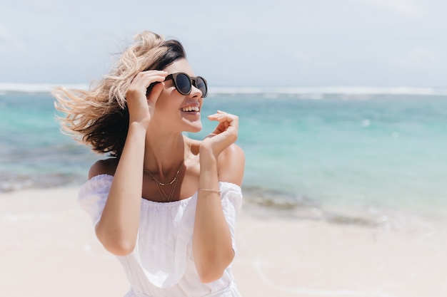 Foto ao ar livre de mulher inspirada sorridente com penteado curto, posando no resort. jocund bronzeada mulher caucasiana rindo da costa do mar.