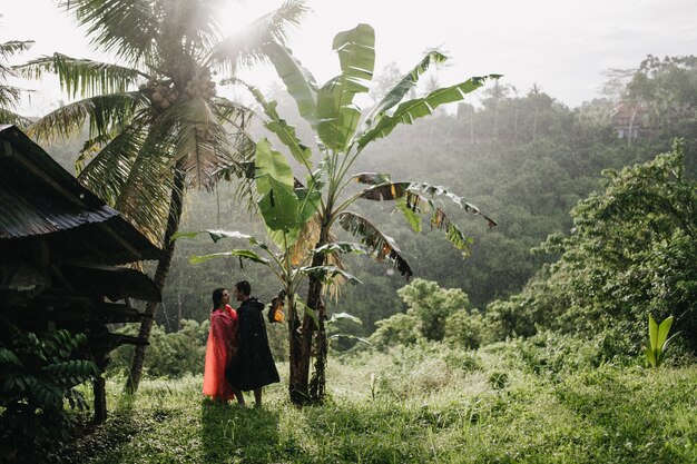 Foto ao ar livre de turista feminina na capa de chuva rosa, posando com o namorado na selva. Retrato de casal relaxando na floresta tropical.