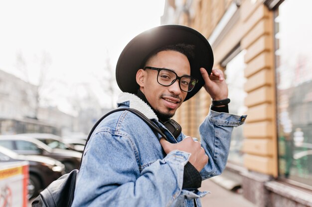 Foto ao ar livre de um cara moreno na moda com pele castanha clara. Foto de um homem Africano despreocupado em traje jeans, relaxando na rua.