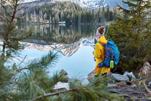 Foto ao ar livre de um jovem viajante com uma bolsa, de costas para a câmera, apreciando montanhas, ar puro e um pequeno lago