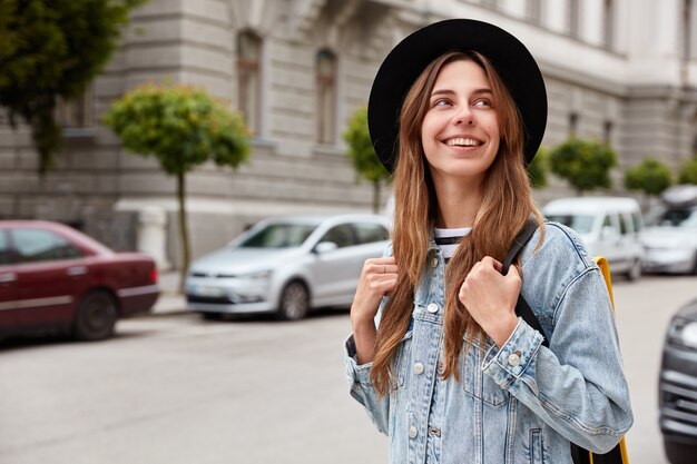Foto ao ar livre de uma bela mulher europeia passeando pela cidade, passando o tempo livre, recriando durante as férias, usando chapéu e jaqueta jeans