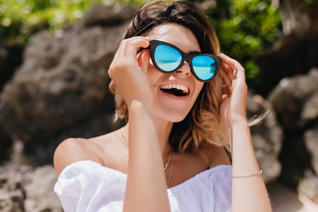 Foto ao ar livre de uma linda mulher europeia olhando em volta com um sorriso. Mulher feliz em óculos de sol se divertindo no resort.