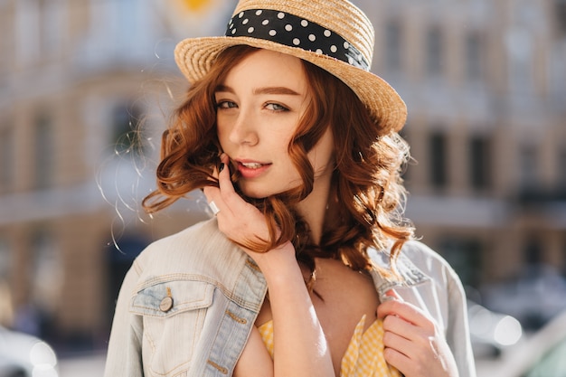 Foto ao ar livre de uma linda senhora caucasiana de cabelo vermelho, andando pela cidade. Modelo feminino elegante com chapéu de palha, posando com prazer em um dia quente.
