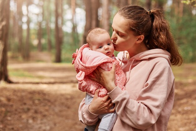 Foto ao ar livre de uma mãe jovem e amorosa, segurando a garotinha nas mãos e beijando-a