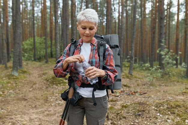 Foto ao ar livre de uma mulher caucasiana de meia-idade ativa carregando uma mochila, abrindo uma garrafa de água, se refrescando durante uma longa e exaustiva caminhada no parque nacional, em pé contra pinheiros