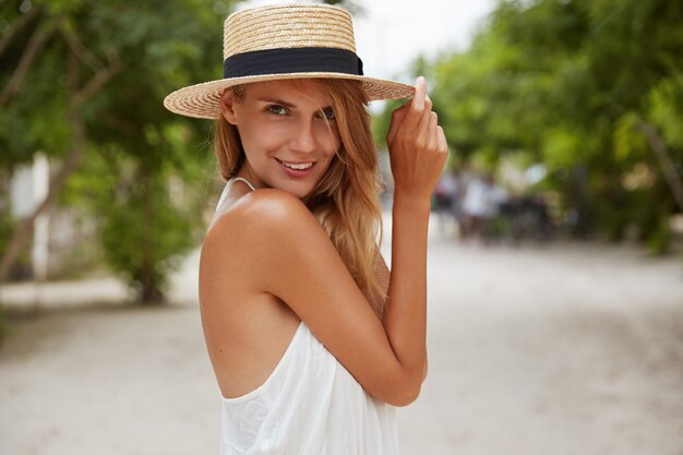 Foto ao ar livre de uma mulher de aparência agradável com pele bronzeada saudável, vestida com um vestido branco e chapéu de verão, posa no parque com uma expressão de satisfação confiante