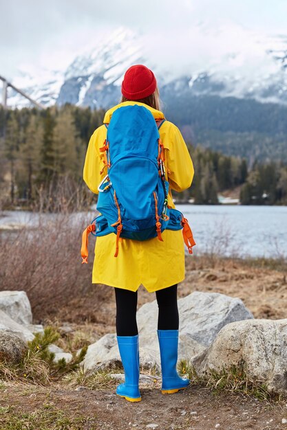 Foto ao ar livre vertical de uma turista admirando a água turquesa do lago, perto de rochas, olhando para montanhas de neve, respirando ar fresco e usando chapéu vermelho