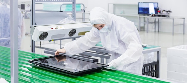 Foto aproximada da linha de montagem verde onde operário em traje de laboratório branco e máscara facial está produzindo um aparelho de tv com alguns equipamentos modernos