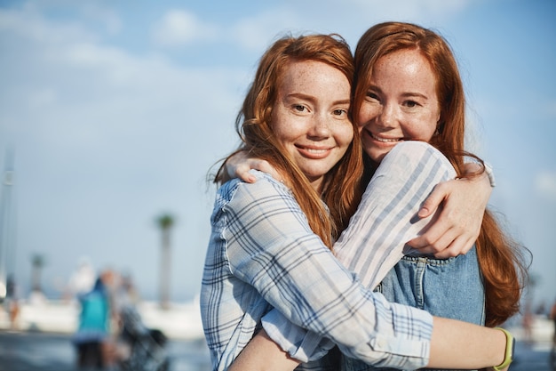 Foto bonita de duas lindas amigas com cabelo ruivo e sardas, se abraçando na rua e sorrindo amplamente, expressando cuidado e amor. Estilo de vida e conceito de relacionamento