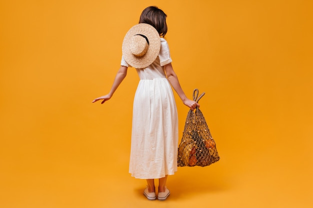Foto completa de garota de vestido midi com chapéu de palha e saco de barbante com frutas em fundo laranja.