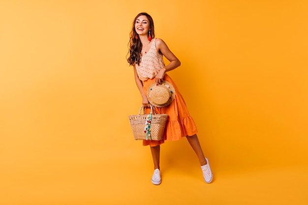Foto completa de mulher inspirada com acessórios de verão. Feliz modelo feminino gengibre em saia laranja segurando o chapéu e a bolsa.