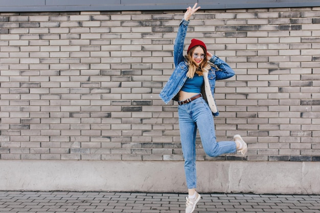 Foto grátis foto completa de senhora encantadora em sapatos esportivos e jaqueta jeans dançando perto da parede de tijolos foto ao ar livre de garota caucasiana pulando na rua com expressão de rosto feliz