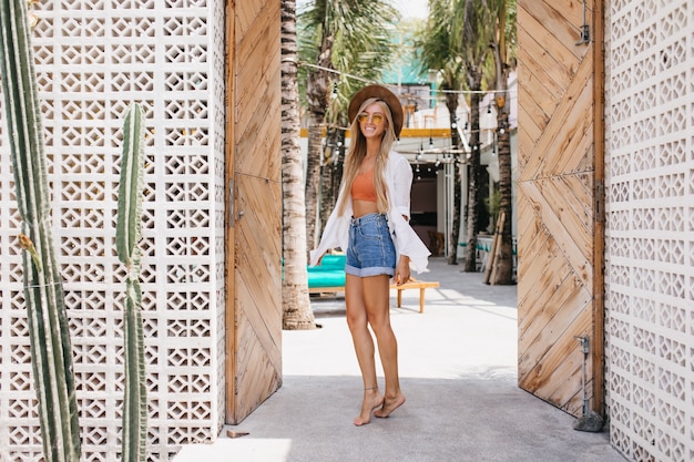 Foto completa de uma garota adorável com um sorriso fofo brincando no resort. Retrato ao ar livre da despreocupada senhora de cabelos louros em shorts jeans, dançando num dia de verão.