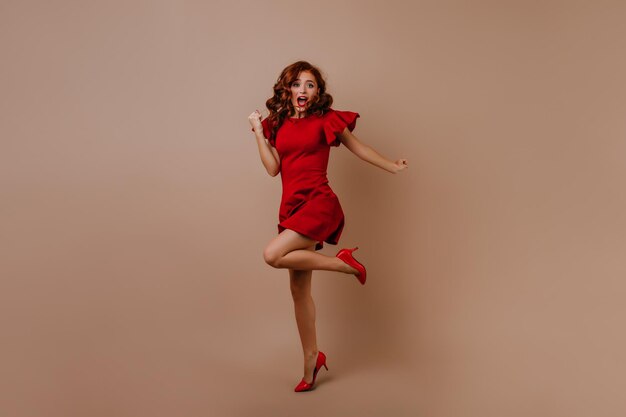 Foto completa de uma mulher caucasiana surpresa com roupa de festa Garota ruiva deslumbrante de vestido vermelho expressando felicidade