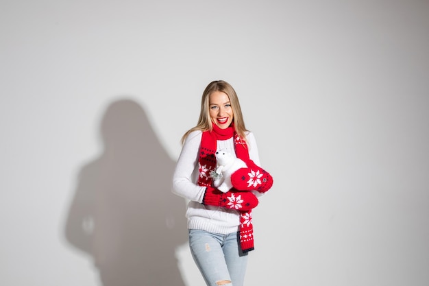 Foto conservada em estoque da mulher branca bonita loira em luvas e cachecol e suéter branco segurando o urso de brinquedo branco bonito e sorrindo.