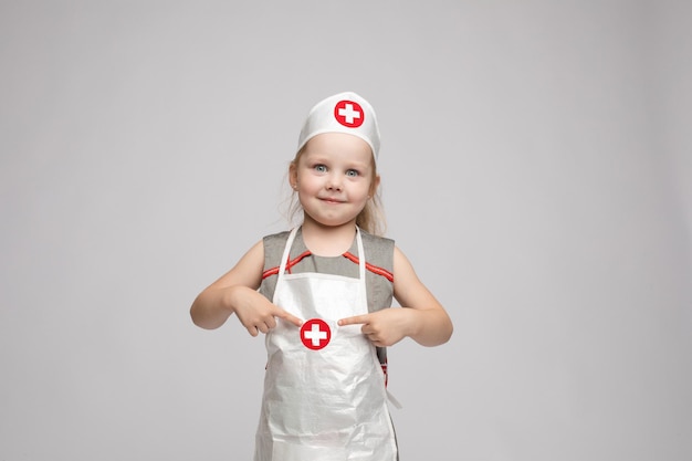 Foto conservada em estoque de linda garotinha de avental branco e um chapéu brincando de médico Ela está apontando para o chapéu com uma cruz branca no círculo vermelho Ela é médica