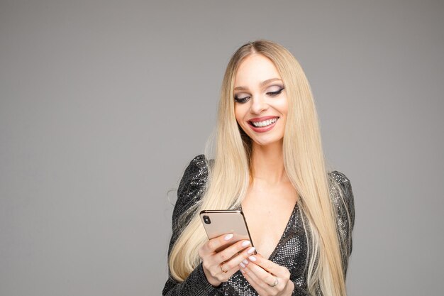 Foto conservada em estoque de uma linda mulher loira com cabelos longos, em um vestido de cocktail cinza navegando no celular, se divertindo e sorrindo.