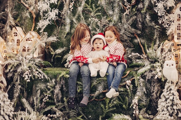 Foto conservada em estoque do adorável garotinho com chapéu de Papai Noel, abraçado por suas irmãs mais velhas em suéteres vermelhos e brancos com padrão de inverno. Crianças com bebê sentado no interior do Natal.