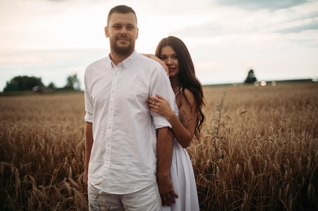 Foto conservada em estoque retrato de namorado barbudo abraçando sua linda namorada tanto em roupas brancas, abraçando no campo de trigo. belo campo de trigo em segundo plano.