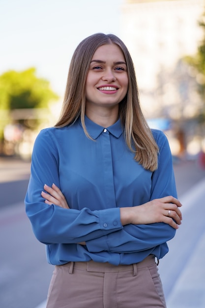 Foto da bela jovem empresária vestindo camisa de chifon azul em pé na rua com os braços cruzados.