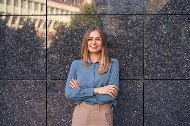 Foto da bela jovem empresária vestindo uma camisa de chiffon azul, de pé com os braços cruzados na parede de mármore cinza