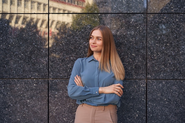 Foto da bela jovem empresária vestindo uma camisa de chiffon azul, de pé com os braços cruzados na parede de mármore cinza