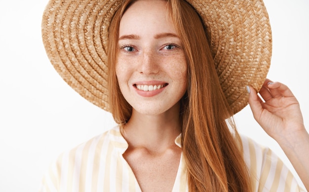 Foto da cabeça de uma garota ruiva encantadora feliz curtindo as férias, sorrindo, segurando um chapéu de palha na cabeça e olhando com uma expressão amigável e encantada