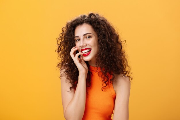 Foto da cintura da encantadora mulher feminina, boba e despreocupada com penteado encaracolado no dedo superior cortado mordendo e sorrindo sensualmente e glamour para a câmera, parecendo sexy e ousada sobre fundo laranja.