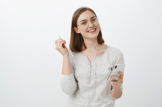 Foto da cintura de uma alegre e encantadora mulher caucasiana de óculos da moda segurando um smartphone, tirando os fones de ouvido e olhando a música favorita de ouvir despreocupada, interrompendo para falar com um amigo