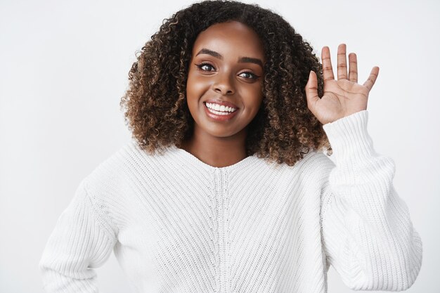 Foto da cintura de uma encantadora mulher de pele escura otimista, de aparência amigável, renunciando à palma da mão erguida em um "olá" ou um gesto de saudação sorrindo abertamente dizendo olá encontrando amigos em um café sobre uma parede branca