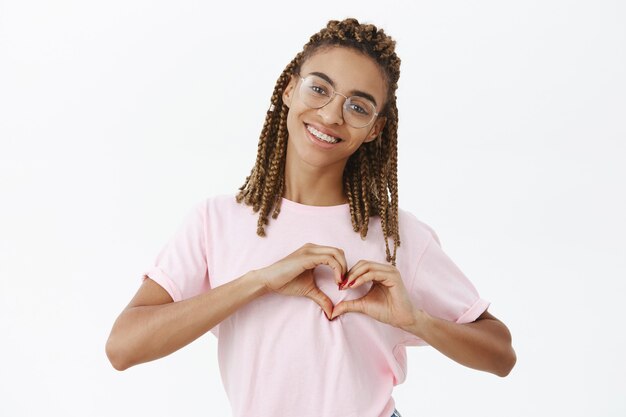 Foto da cintura de uma linda namorada afro-americana atraente e amigável com dreads em uma camiseta rosa mostrando um gesto de coração