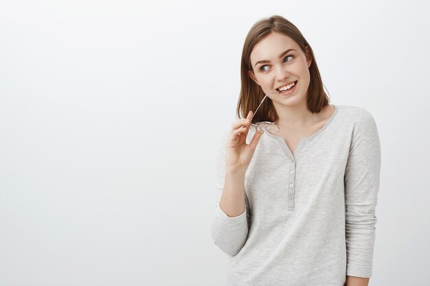 Foto da cintura de uma mulher europeia criativa, cheia de energia e feliz com um corte de cabelo castanho curto, mordendo a borda dos óculos, segurando os óculos na cabeça, inclinando a cabeça de interesse, olhando encantada e intrigada para a esquerda