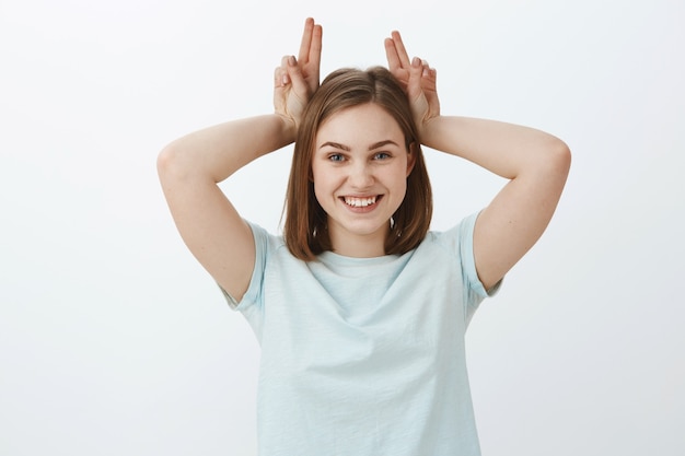 Foto da cintura de uma mulher fofa, enérgica e brincalhona, emotiva em uma camiseta da moda, segurando os dedos na cabeça como se imitasse orelhas de coelho sorrindo alegremente, se divertindo e se divertindo sobre uma parede cinza