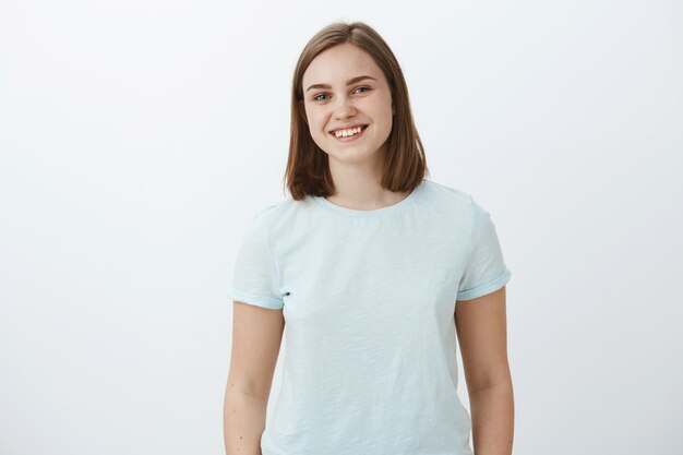 Foto da cintura para cima da ambiciosa, feliz e fofa morena feminina em uma camiseta da moda, sorrindo alegremente, deliciada e satisfeita ao receber os parabéns por ganhar o prêmio sobre a parede branca