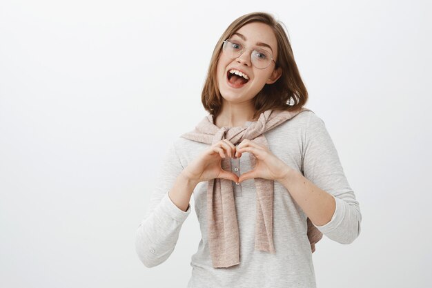 Foto da cintura para cima da sociável, feliz e amigável morena caucasiana com óculos da moda, blusa e pulôver amarrados no pescoço mostrando um gesto de coração confessando estar apaixonado com um sorriso largo e cabeça inclinada