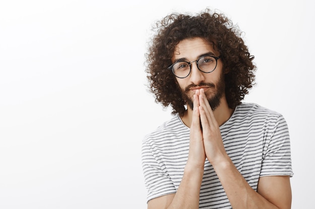 Foto da cintura para cima de um modelo masculino hispânico sonhador bonito com barba e penteado afro, segurando as mãos em oração perto da boca e olhando pensativamente